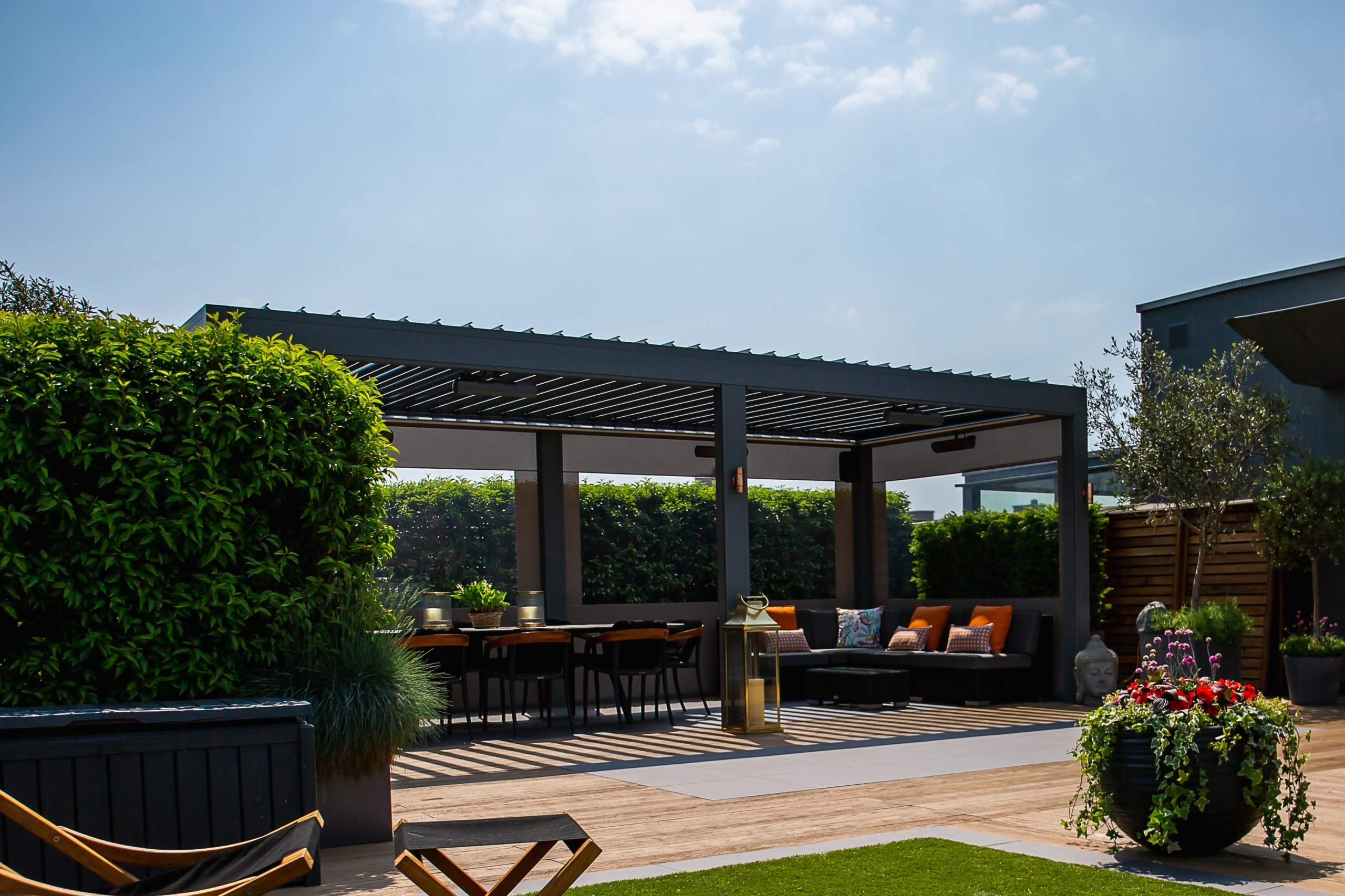 A pergola with seating and a dining area is set on a wooden deck; surrounded by lush greenery and planters under a clear blue sky.