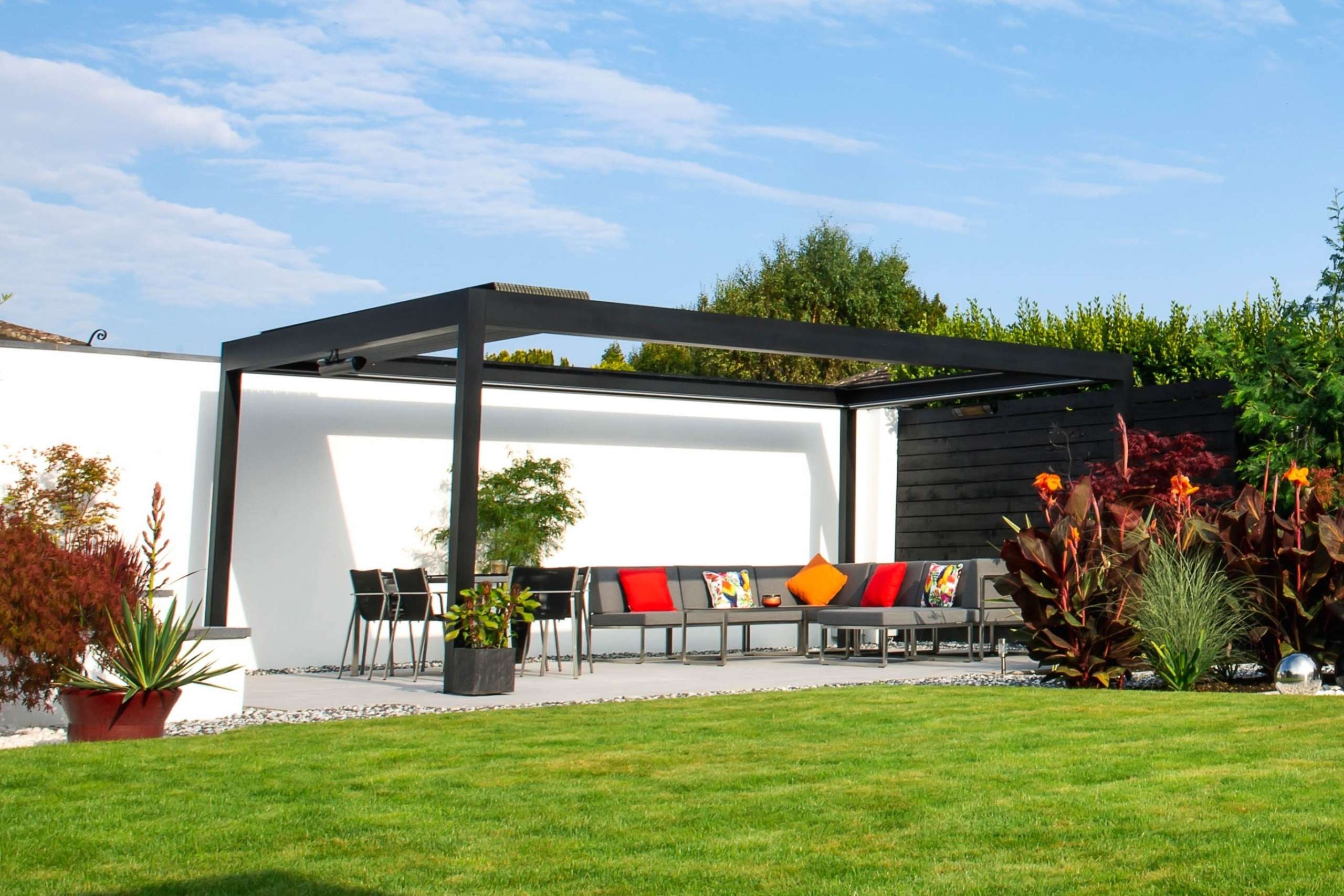 A louvered grey aluminium pergola in front of a white wall on a blue sunny day. There is vibrant green grass in the foreground.