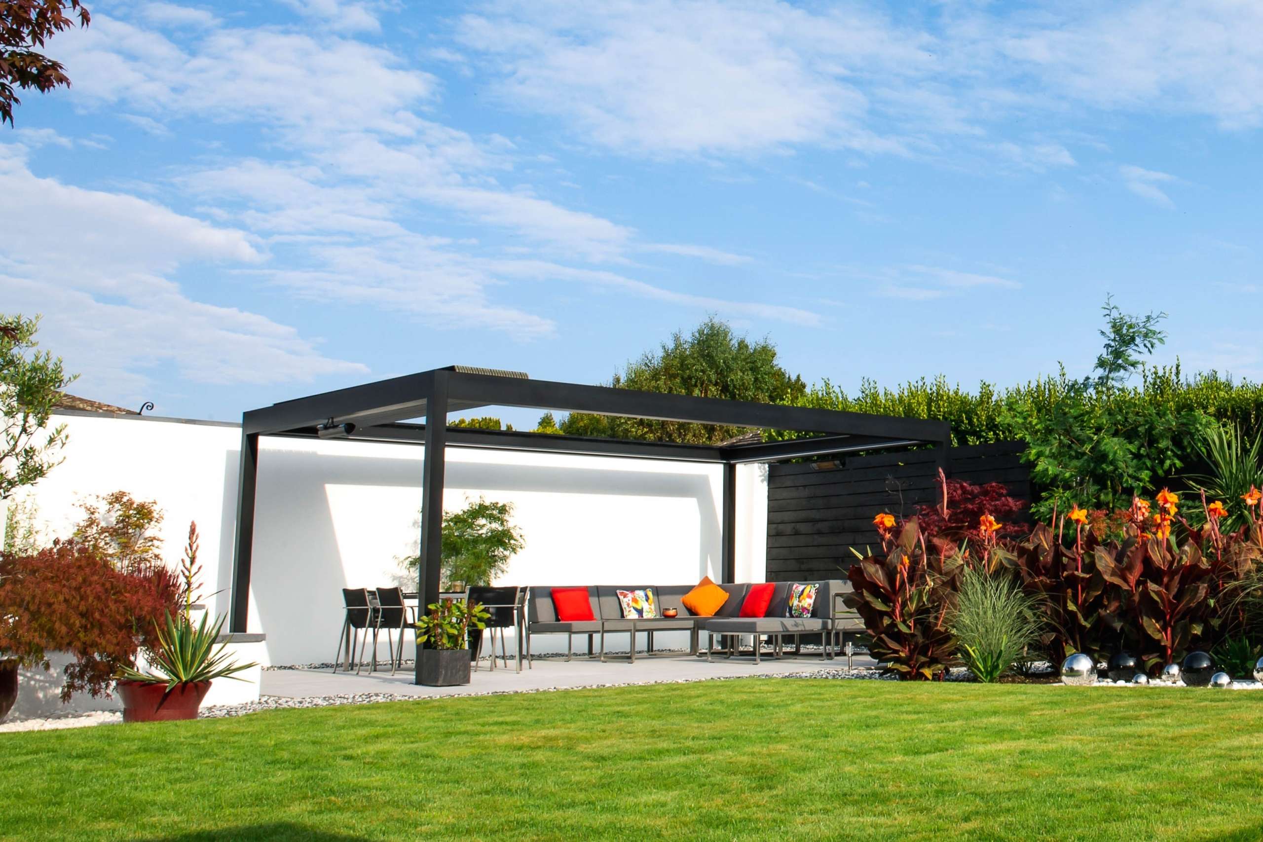 A louvered grey aluminium pergola in front of a white wall on a blue sunny day. There is vibrant green grass in the foreground.