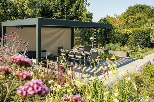 A modern pergola shelters a dining table and chairs, surrounded by lush, colorful garden plants, with a wooden building to the right and dense greenery in the background.