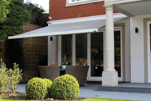 Awning extends over wicker patio furniture on a stone terrace, surrounded by manicured shrubs and trees, adjacent to a brick house with large glass doors.