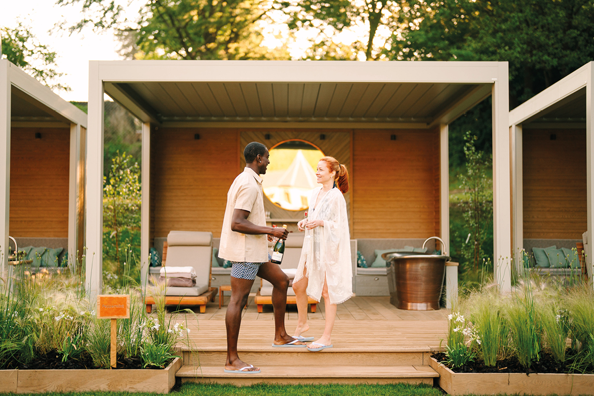 A man and woman converse on a wooden deck under a modern pavilion. They wear casual summer attire, surrounded by greenery and lounge furniture, with warm sunlight filtering through trees.