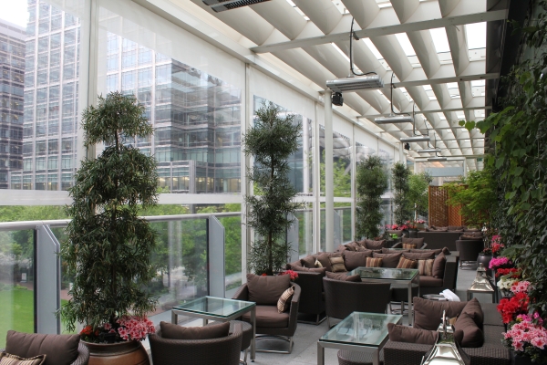 Wicker chairs and glass tables arranged in a spacious, modern outdoor terrace, surrounded by lush greenery and colorful flowers, with tall glass walls offering views of adjacent buildings.