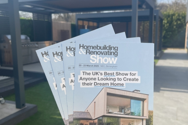 Brochures depicting a modern house are held in an outdoor area with neatly maintained grass, patio seating, and a pergola. Text: "Homebuilding & Renovating Show. 20 - 23 March 2025, NEC, Birmingham. The UK’s Best Show for Anyone Looking to Create their Dream Home."